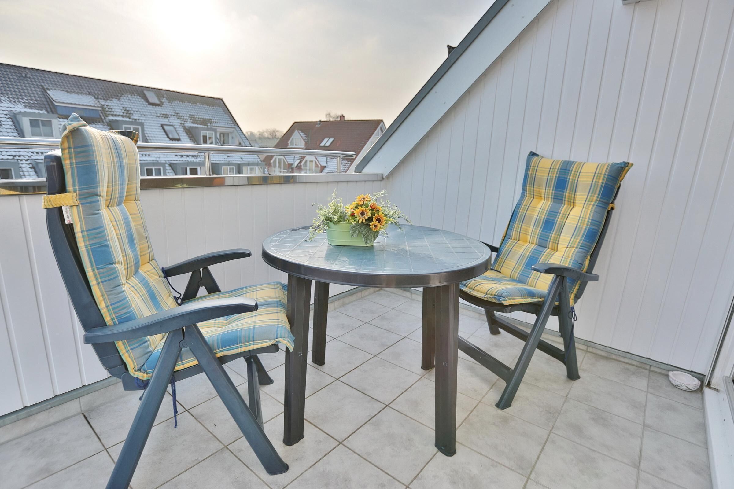 A balcony with two chairs and a table, a vase with flowers, and a view of buildings in the background.