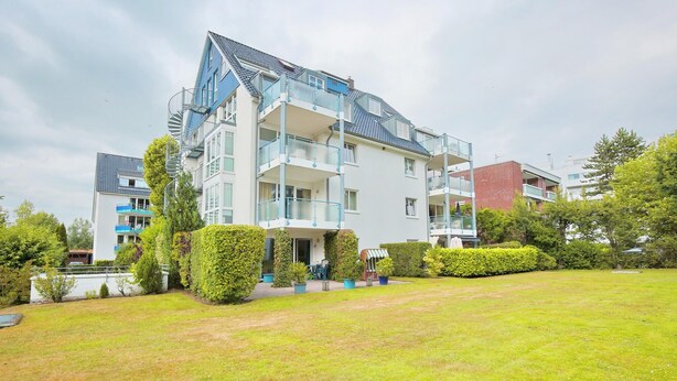 A modern multi-story residential building with balconies, a well-maintained lawn, and surrounding greenery.