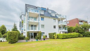 A multi-story residential building with balconies, a grassy front yard, and potted plants.