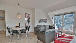 A modern living room with a dining area, a grey sofa, a red rug, and a large window offering a view of buildings.