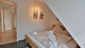A bedroom with a bed, pillows, and a white rug. There are two paintings on the wall and a door leading to another room.