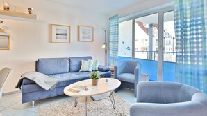 Ein modernes Wohnzimmer mit einem blauen Sofa, einem runden Couchtisch und einem blauen Sessel. An den Wänden hängen Bilderrahmen, auf dem Couchtisch steht ein Topf mit einer Pflanze und durch große Fenster ist ein Blick auf Gebäude möglich.