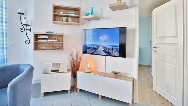 Ein modernes Wohnzimmer mit einem Flachbildfernseher, Holzregalen, einem weißen Konsolentisch, einem blauen Sessel und einer Tür zu einem anderen Raum.