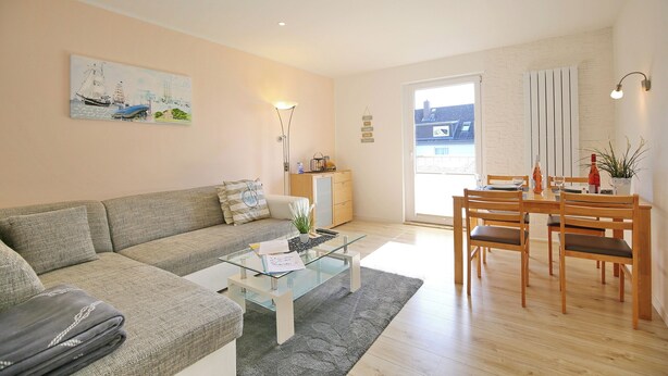 A living room with a sofa, coffee table, dining table with chairs, and a painting on the wall. - Ferienhaus Brandt Appartement 3 Möwenkieker (Haffkrug)