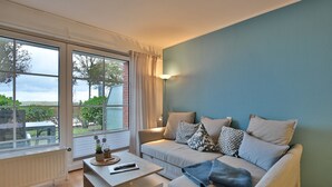 A modern living room with a sofa, coffee table, and large windows. - Windrose Appartement 1 und 3 (Scharbeutz)