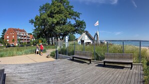 A wooden boardwalk with benches, a flag, and a building in the background. - Windrose Appartement 1 und 3 (Scharbeutz)