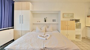 A hotel room with a bed, a wardrobe, a small shelf with decorative items, and a kitchenette area. - Windrose Appartement 1 und 3 (Scharbeutz)