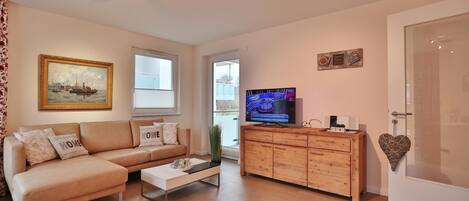 A modern living room with a beige sofa, a coffee table, a TV on a wooden cabinet, and a door with a heart-shaped decoration.