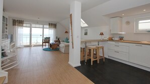 A spacious living area with a large window, a dining area with a table and chairs, and a kitchen with white cabinets and a wooden countertop. - Meerglück Appartement 5 (Haffkrug)