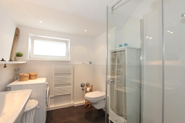 Un baño moderno con cabina de ducha de vidrio, inodoro blanco, lavamanos empotrado y ventana que proporciona luz natural.