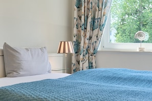 A bedroom with a bed, a lamp, a window with curtains, and a view of trees. - Dünenhaus 3 Appartement 7 (Scharbeutz)