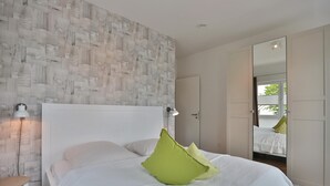A bedroom with a white bed, green pillows, a white nightstand, a white lamp, and a wooden wall. - Haus Meeresbrise Appartement 18 (Haffkrug)