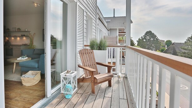 A wooden deck with a white railing, a wooden chair, and a glass door leading to an interior space with a blue sofa and a small table. - Haus Meeresbrise Appartement 18 (Haffkrug)