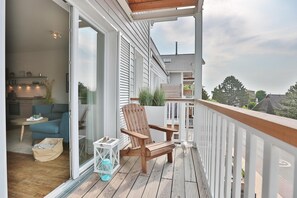 A wooden deck with a white railing, a wooden chair, and a glass door leading to an interior space with a blue sofa and a small table. - Haus Meeresbrise Appartement 18 (Haffkrug)