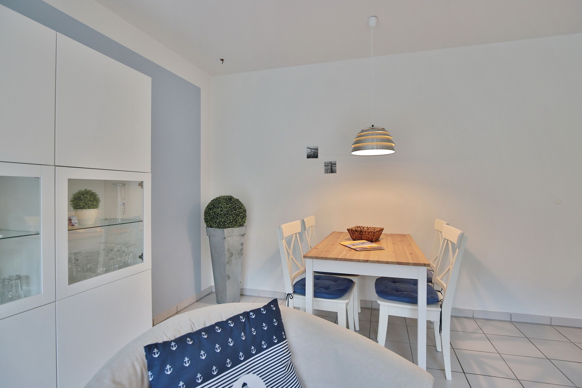 Una cocina moderna con muebles blancos, una mesa de comedor con sillas, una lámpara colgante y una planta en maceta.