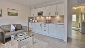 A modern kitchen with white cabinets, a microwave, and a sink. There is a glass coffee table in the living area with a sofa and a painting on the wall. - Haus Hansastrasse Appartement 2 (Scharbeutz)