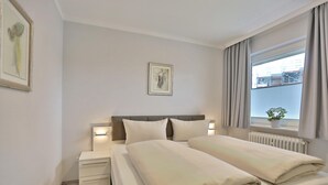 A hotel room with a double bed, bedside tables, a nightstand, a framed picture on the wall, and a window with curtains. - Haus Hansastrasse Appartement 2 (Scharbeutz)