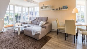 En moderne stue med sofa, sofabord, spisebord med stole, og et stort vindue der giver udsigt over naturen.