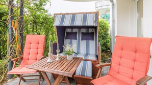 An outdoor seating area with two cushioned chairs and a wooden table, surrounded by greenery and a building in the background. - Ostseeperle Appartement 2 (Scharbeutz)