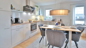 A modern kitchen with white cabinets, a built-in oven, and a dining area with a wooden table and grey chairs. - Ostseeperle Appartement 2 (Scharbeutz)