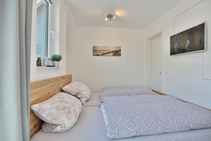 A bedroom with a wooden headboard, two patterned pillows, a bed with a patterned bedspread, a window with a view of the sea, a painting on the wall, and a ceiling light fixture. - Bude 37 Appartement 6 (Scharbeutz)