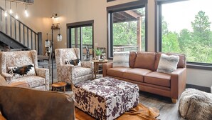 A spacious living room with a large window, a staircase, and a cozy seating area with a leather sofa and ottoman. - New Spacious 3200 sq. ft. Cabin w/ Hot Tub Across Street from Clubhouse Pool (Lead)
