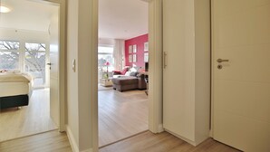 A modern apartment with a wooden floor, a bed in the left room, a living area with a sofa and a table, and a red accent wall. - Strandperle Appartement 203 (Haffkrug)