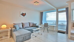 Ein modernes Wohnzimmer mit einem grauen Ecksofa, einem kleinen weißen Couchtisch, einem grauen Sessel und einem großen Fenster mit Blick auf das Meer.