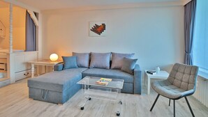 Ein modernes Wohnzimmer mit einem grauen Ecksofa, einem Glastisch, einem grauen Sessel und einem kleinen weißen Beistelltisch mit einer Lampe.