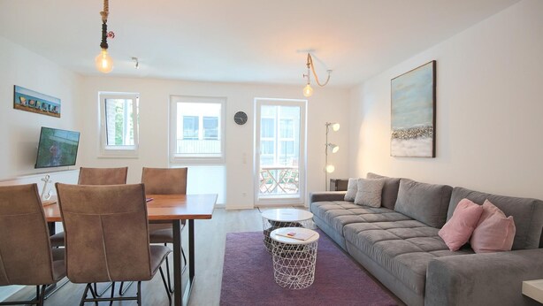 Een woonkamer met een grijze bank, een houten eettafel met stoelen, een paars tapijt en een televisie aan de muur.