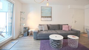 Een moderne woonkamer met een grijze bank, twee ronde witte bijzettafeltjes, een paars vloerkleed en een schilderij van een strand.