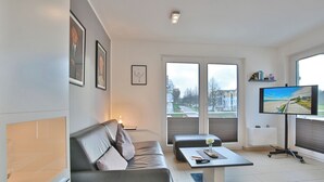 Ein modernes Wohnzimmer mit Sofa, Couchtisch und Fernseher. Große Fenster bieten einen Ausblick nach draußen.