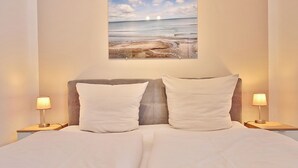 Cama bem arrumada com roupa de cama branca, duas luminárias de cabeceira e uma cena de praia emoldurada na parede.