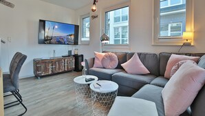 Ett modernt vardagsrum med en grå soffa, vita, runda sidobord, en antik resväska och en platt-TV som är monterad på väggen.