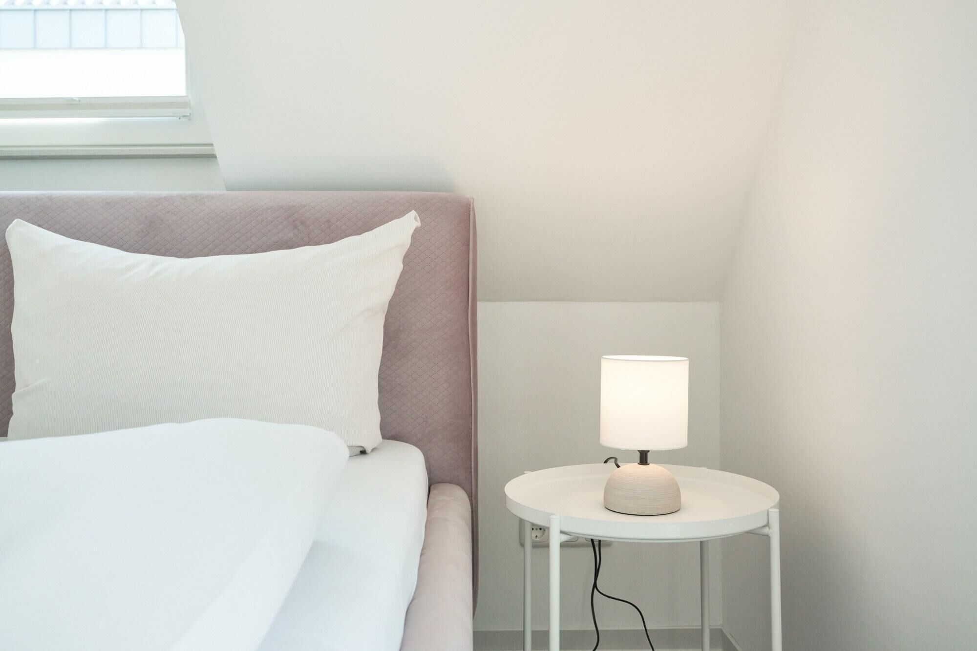 Een opgemaakt bed met wit en roze beddengoed, een bedlampje op een klein wit tafeltje en een raam met jaloezieën.