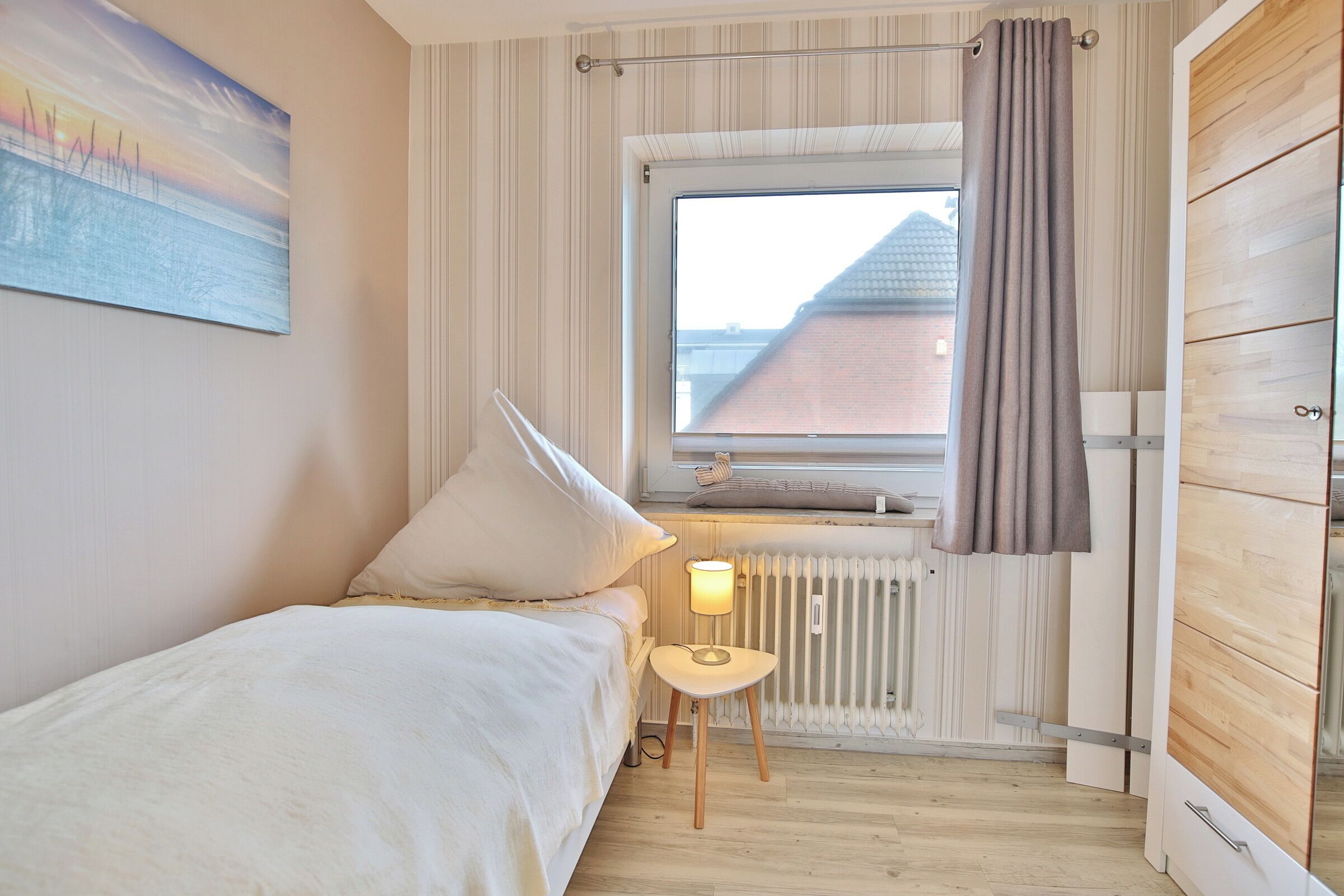 Ein Schlafzimmer mit einem Bett, einem Nachttisch, einem Fenster mit Vorhängen, einem Heizkörper und einem Holzschrank.