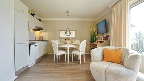 A modern kitchen with white cabinets, a dining table with chairs, a sofa with an orange pillow, and a TV on the wall. - Strandschlösschen Appartement 28 (Haffkrug)