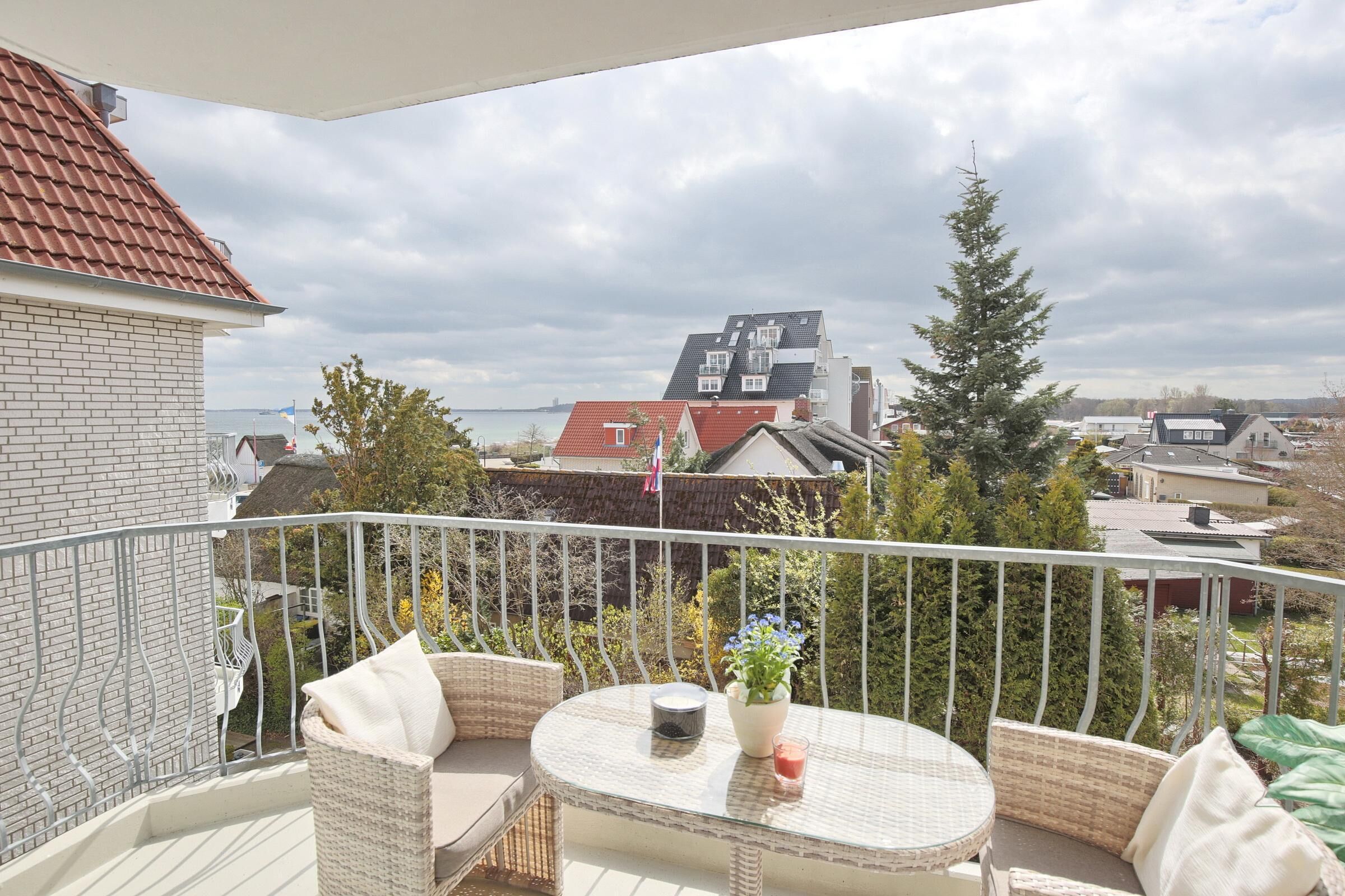 Un balcón con mobiliario de mimbre y una mesa, con vistas a una zona residencial con casas y una bandera.