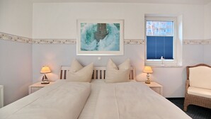 Ein Schlafzimmer mit einem großen Bett, zwei Nachttischen mit Lampen, einem Korbstuhl, einem gerahmten Bild an der Wand und einem Fenster mit Jalousien.