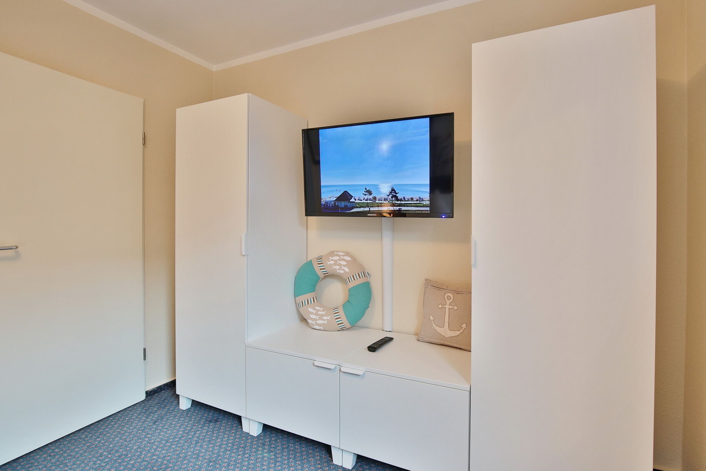 A hotel room with a television mounted on a white cabinet, a remote control on the cabinet, and a decorative anchor pillow.