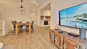 Un apartamento moderno con comedor, cocina y un televisor grande mostrando una escena de playa.