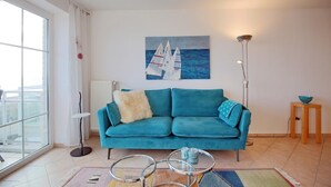 A living room with a teal sofa, a painting of sailboats, a floor lamp, and a small wooden table with a bowl. - Stranddomizil Appartement 14 (Haffkrug)