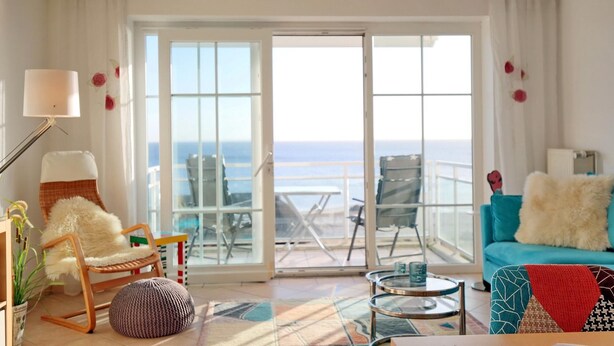 A spacious living room with a large sliding glass door leading to a balcony with a view of the ocean. There is a cozy seating area with a chair and ottoman, a small round table, and a blue sofa with a fluffy throw. - Stranddomizil Appartement 14 (Haffkrug)