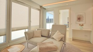 A modern living room with a beige sofa, a matching armchair, and a small side table. There is a large window with blinds, a fireplace with a vase, and a framed picture on the wall. - Villa Seeperle II Herzmuschel Appartement 18 (Scharbeutz)