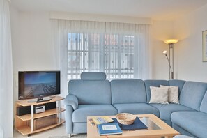 Una sala de estar moderna con un sofá azul, un televisor sobre un mueble de madera, una lámpara de pie y una ventana con cortinas translúcidas.