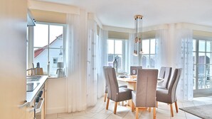 A modern kitchen with a dining area, featuring a large window, white curtains, and a chandelier.