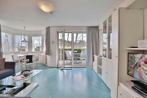 A modern hotel room with a glass coffee table, a sofa, a TV, and a view of the garden through the door.