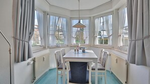 A corner room with a white table, four chairs, a hanging lamp, and large windows with curtains. - Seesternresidenz Appartement 16 (Scharbeutz)