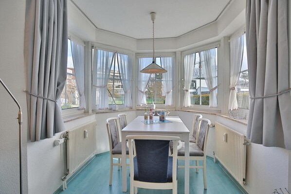 A corner room with a white table, four chairs, a hanging lamp, and large windows with curtains.