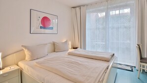 A hotel room with a bed, a bedside table, a nightstand, a chair, and a window with sheer curtains. - Seesternresidenz Appartement 16 (Scharbeutz)